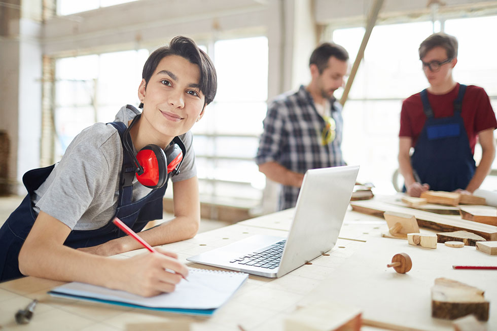 Ausbildungsdokumentation am Arbeitsplatz mit BLok. Azubi mit Laptop und Notizheft in einer Werkstatt.